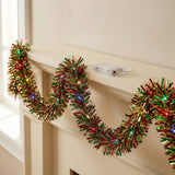 Lighted red, green, and gold tinsel garland hangs in loops along a cream mantel, with a clear battery box resting above.