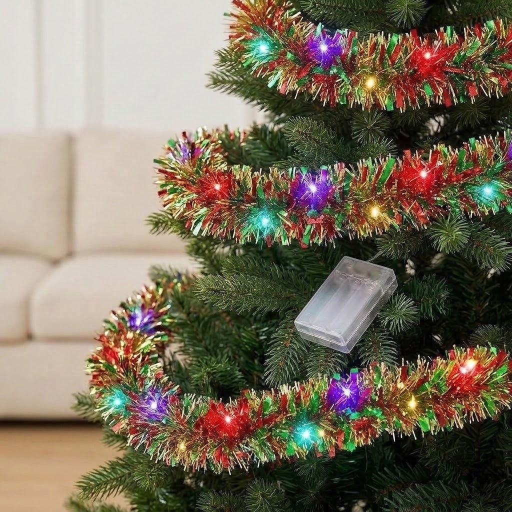A red, green, and gold tinsel lighted garland glows around Christmas tree branches, with a clear battery box resting among the needles.