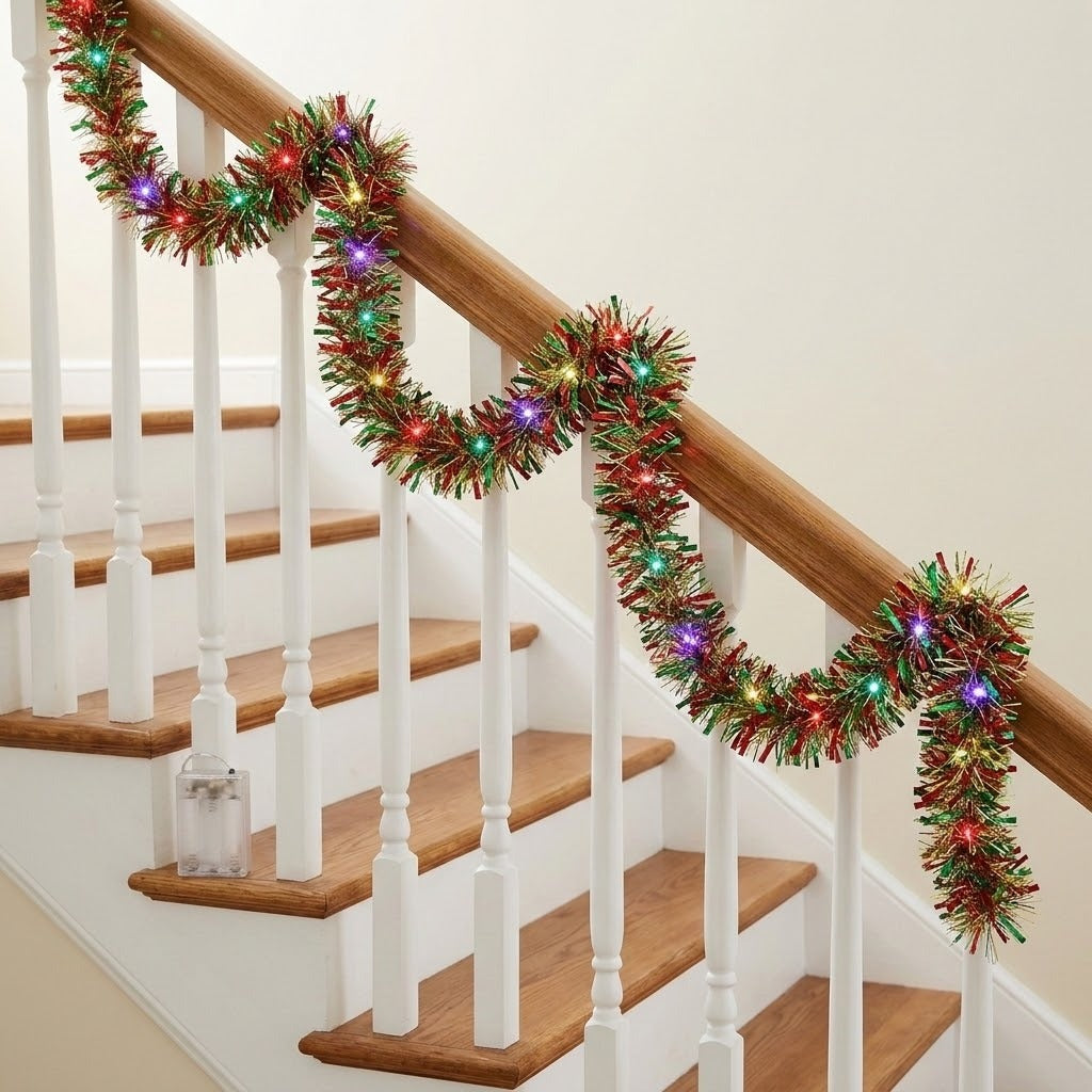 Lighted red, green, and gold tinsel garland drapes along a wooden stair rail above white spindles, with a battery box on one step.