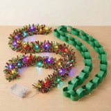 Red, green, and gold tinsel lighted garland with multicolor lights and a clear battery box beside a green paper chain on light wood.