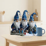 Three blue-hatted, white-bearded Christmas gnomes printed on a clear tabletop decoration stand beside a blue mug on a light wooden table.