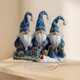 Acrylic tabletop decoration featuring three blue-hatted Christmas gnomes stands on a clear oval base on a wooden shelf against beige walls.