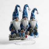 Angled acrylic tabletop decoration shows three blue-hatted gnomes, printed gifts, and snowy branches above a clear oval base against white.