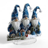 Acrylic tabletop decoration depicting three blue-hatted Christmas gnomes and gifts rests on a clear base against a white background.