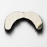Black costume facial hair lies face down on white, with cream release paper partially peeled from the mustache backing at one corner.