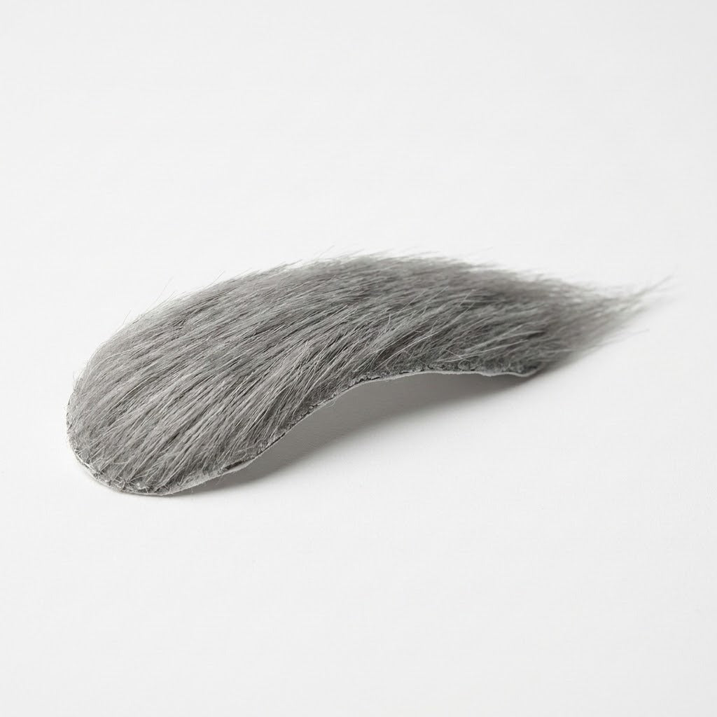 A gray costume facial hair eyebrow rests on a white surface, showing fine strands and a thin backing edge.