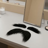 Black costume facial hair, comprising a mustache and two eyebrows, lies on a white vanity beside a tabletop mirror and makeup.