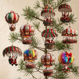Christmas ornaments with red, green, cream, and rainbow Santa balloon and parachute designs hang from sparse pine branches against beige.
