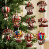 Colourful Santa balloon and parachute Christmas ornaments hang beside dense evergreen branches with warm blurred lights against a cream background.