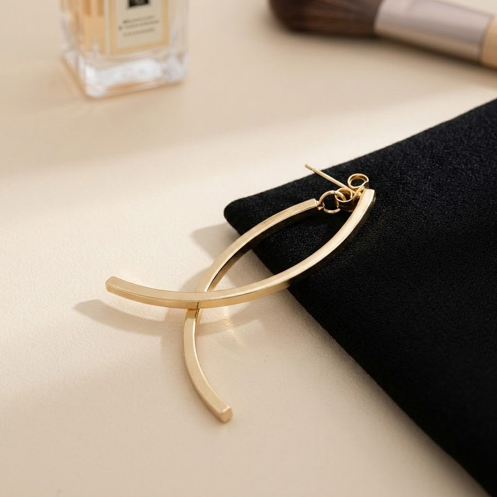 A gold-tone drop earring with crossing curved bars rests partly on black fabric beside a blurred perfume bottle and makeup brush.