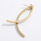 A gold-tone drop earring lies diagonally on white, with crossing curved bars, a connecting ring, and butterfly backing visible.