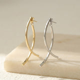 Gold-tone and silver-tone drop earrings with crossing curved bars stand on a cream block against a beige background.