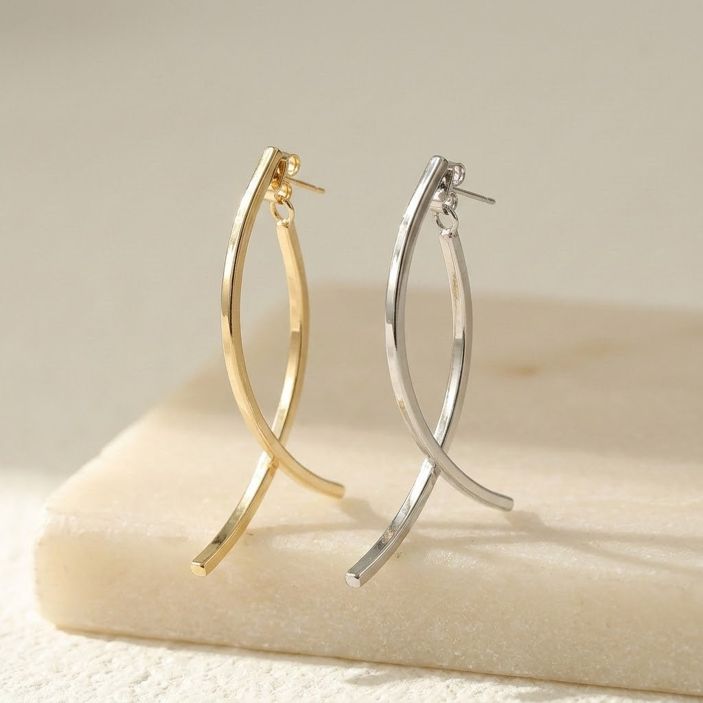 Gold-tone and silver-tone drop earrings with crossing curved bars stand on a cream block against a beige background.