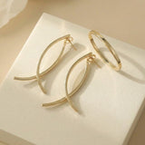 Two gold-tone drop earrings with crossing curved bars rest beside a slim bangle on a textured cream block in warm light.