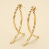 A pair of polished gold-tone drop earrings with crossing curved bars and visible stud posts stands against a cream background.