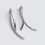 A pair of polished silver-tone drop earrings with long, crossing curved bars rests on a pale gray background.