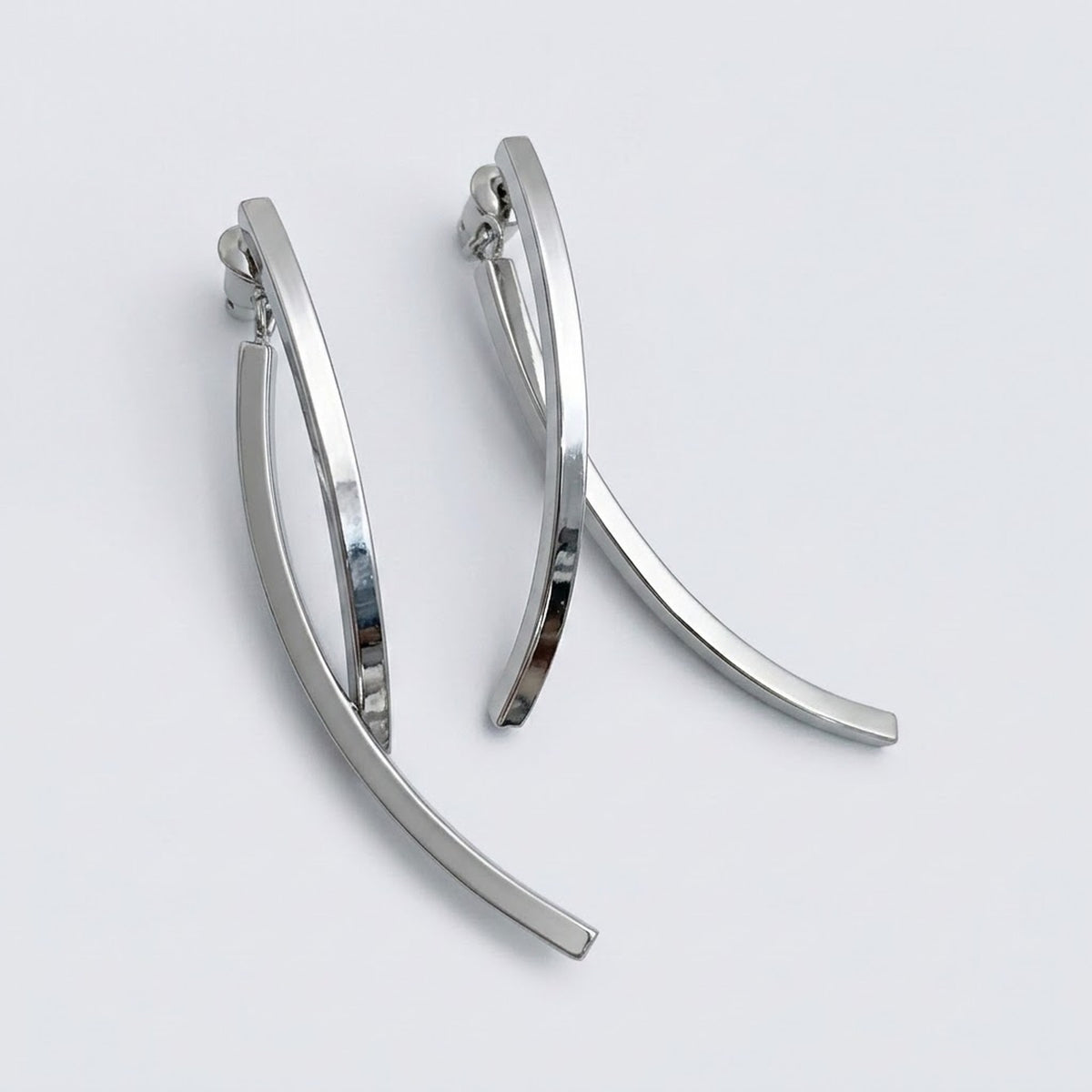 A pair of polished silver-tone drop earrings with long, crossing curved bars rests on a pale gray background.