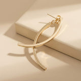 A gold-tone drop earring with crossing curved bars rests across a raised cream block, casting curved shadows in warm light.