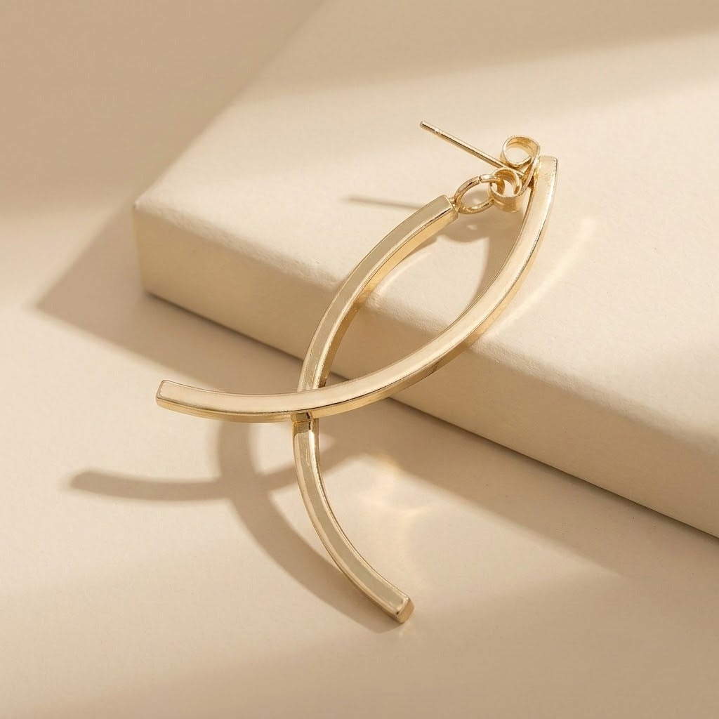 A gold-tone drop earring with crossing curved bars rests across a raised cream block, casting curved shadows in warm light.