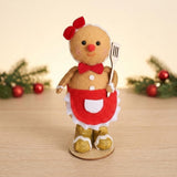 Tan gingerbread sitter with red bows, red apron, and gold boots stands on a wooden tabletop before greenery and red ornaments.