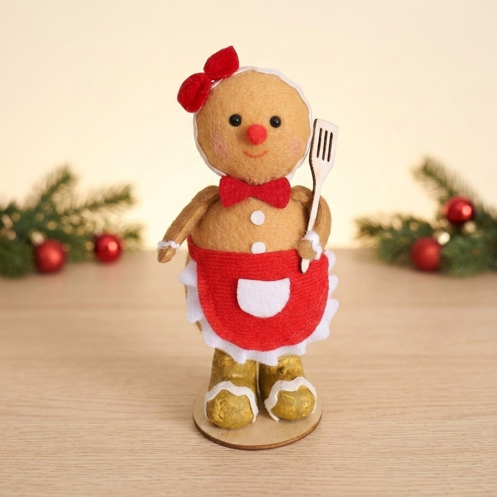 Tan gingerbread sitter with red bows, red apron, and gold boots stands on a wooden tabletop before greenery and red ornaments.