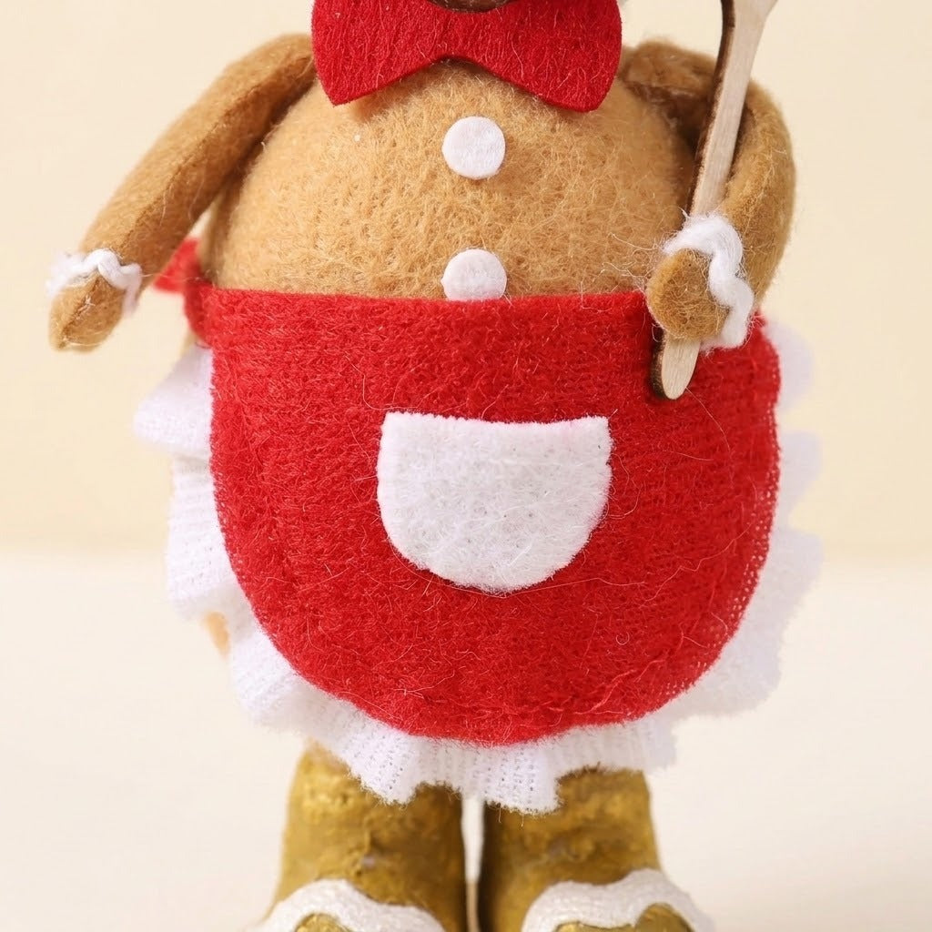 Close-up shows the tan plush gingerbread sitter’s red apron, white pocket, ruffled trim, white buttons, and spatula against a cream background.
