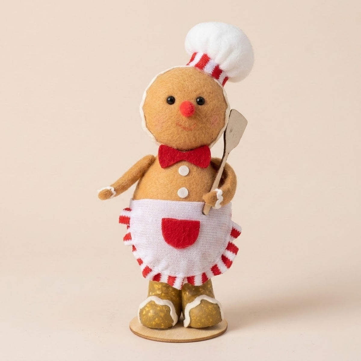 Tan plush gingerbread sitter wears a white chef hat, red bow tie, and white apron while holding a spatula against a beige background.