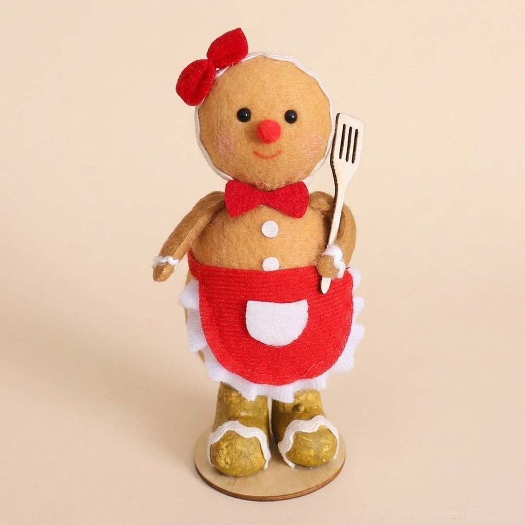 Tan plush gingerbread sitter with a red hair bow, red apron, and gold boots holds a slotted spatula against a beige background.