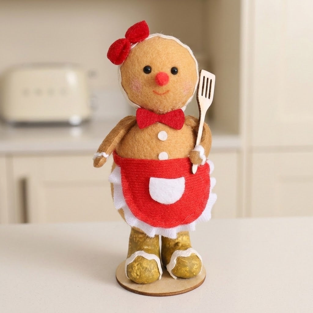 Tan plush gingerbread sitter wears a red apron and gold boots on a white kitchen counter with a blurred toaster behind.