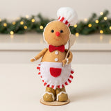 Tan plush gingerbread sitter wears a white chef hat and apron on a white surface before greenery and warm lights.
