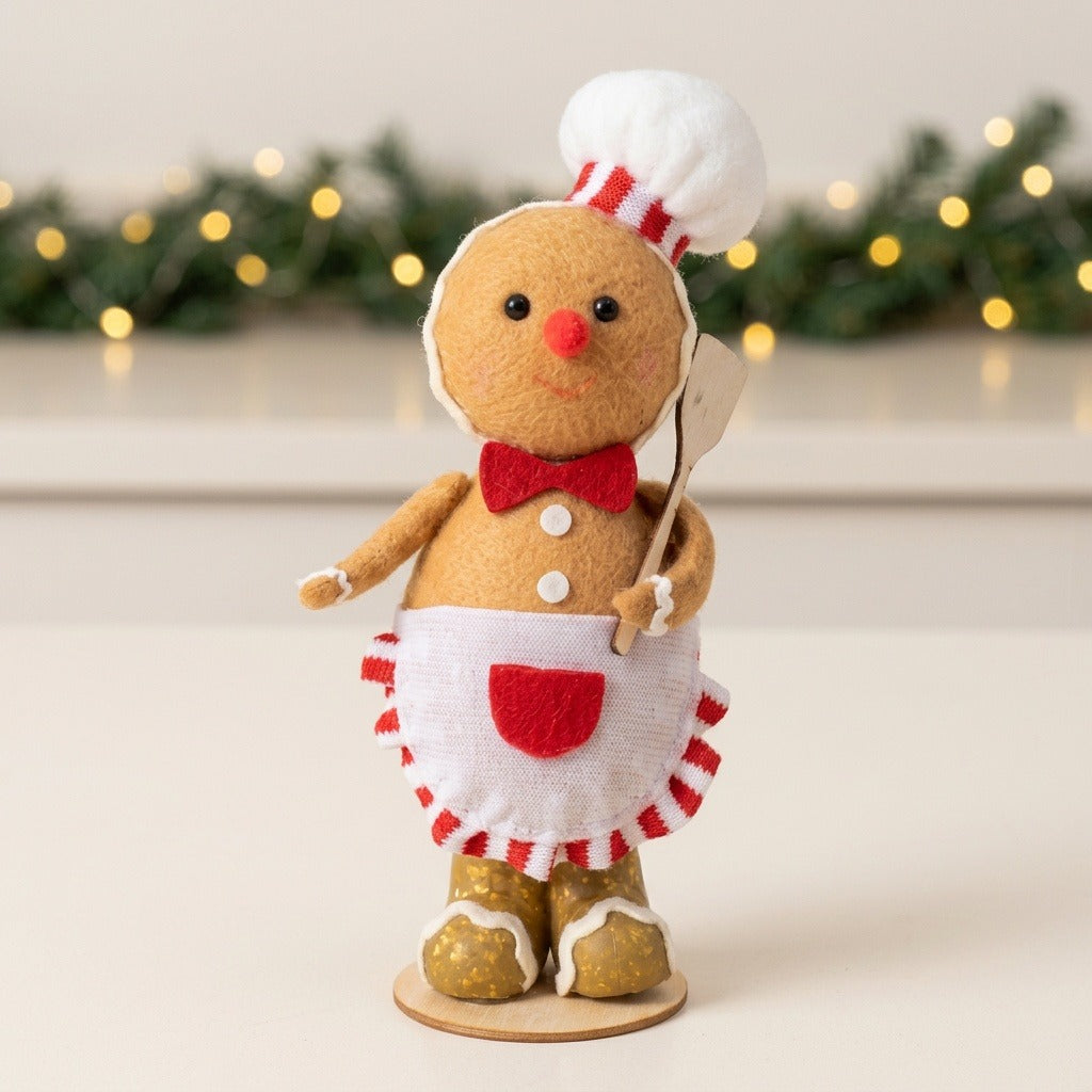 Tan plush gingerbread sitter wears a white chef hat and apron on a white surface before greenery and warm lights.