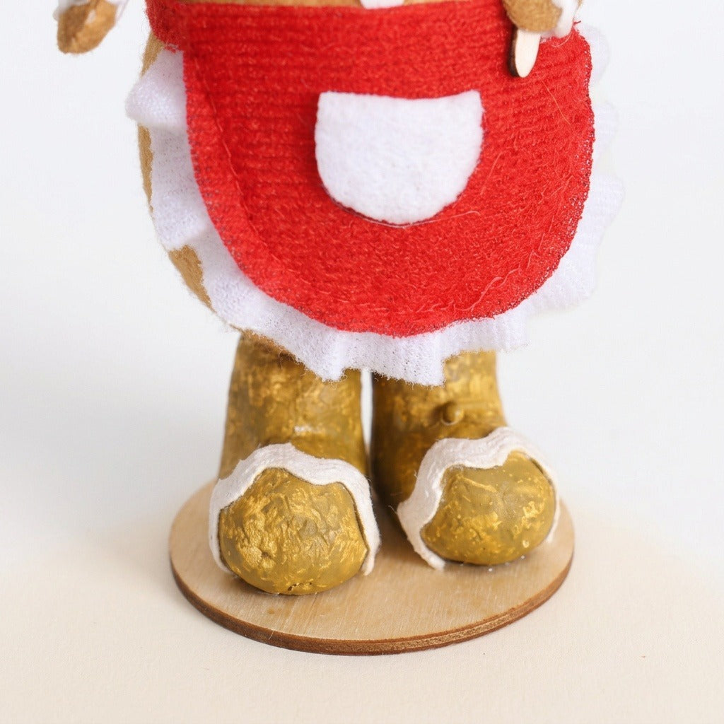 Close-up of the gingerbread sitter shows a red fabric apron, white pocket and ruffled trim, gold boots, and round wooden base.