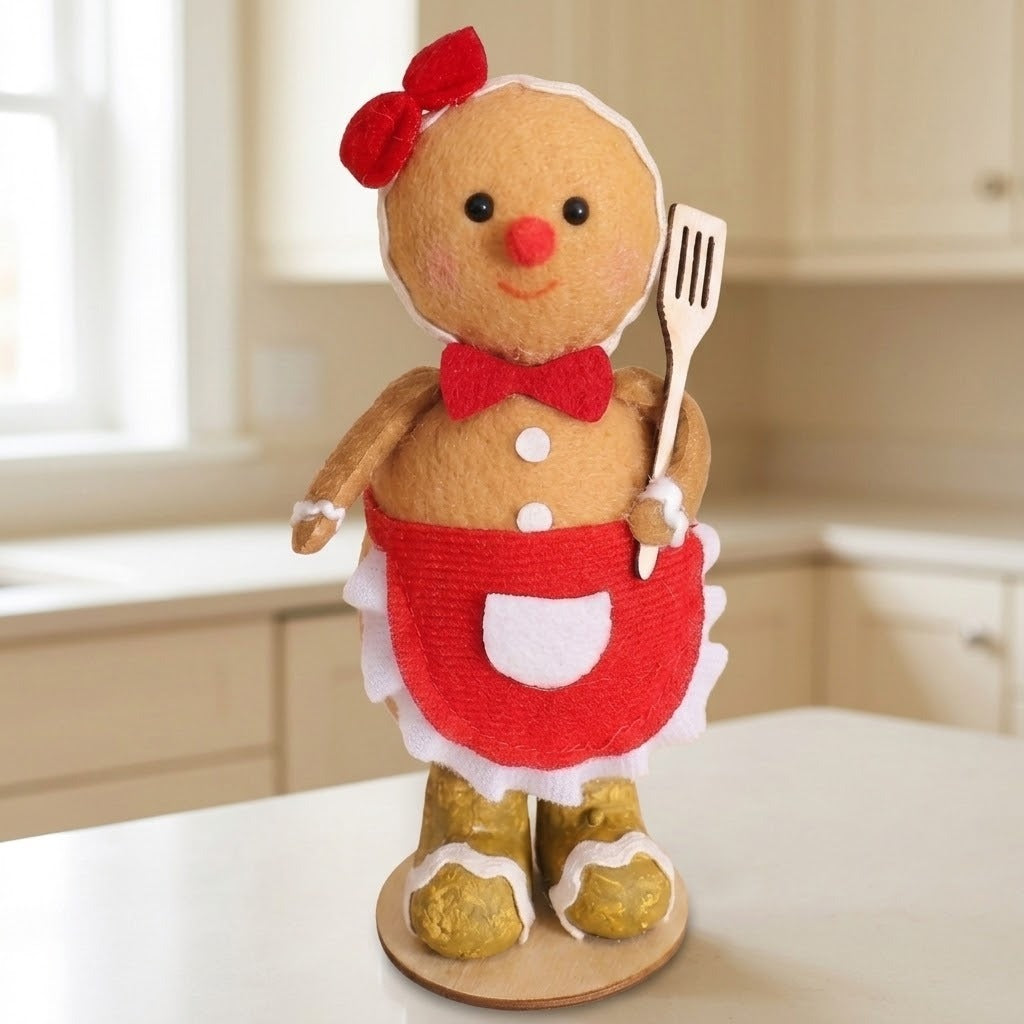 Tan plush gingerbread sitter with a red hair bow and apron holds a slotted spatula on a white kitchen countertop.