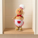 Tan plush gingerbread sitter wears a white chef hat and apron while holding a spatula on a wooden shelf between cream cabinets.