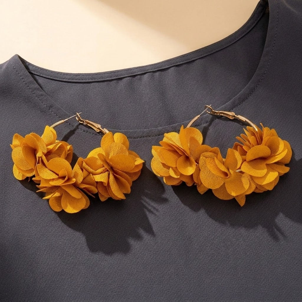 A pair of chunky gold-tone hoop earrings decorated with layered mustard yellow fabric flowers, displayed on a dark gray top against a light beige background.