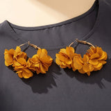 A pair of chunky gold-tone hoop earrings decorated with layered mustard yellow fabric flowers, displayed on a dark gray top against a light beige background.