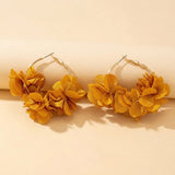 A pair of large gold-tone hoop earrings adorned with clusters of layered mustard yellow fabric flowers, displayed on a warm beige background.
