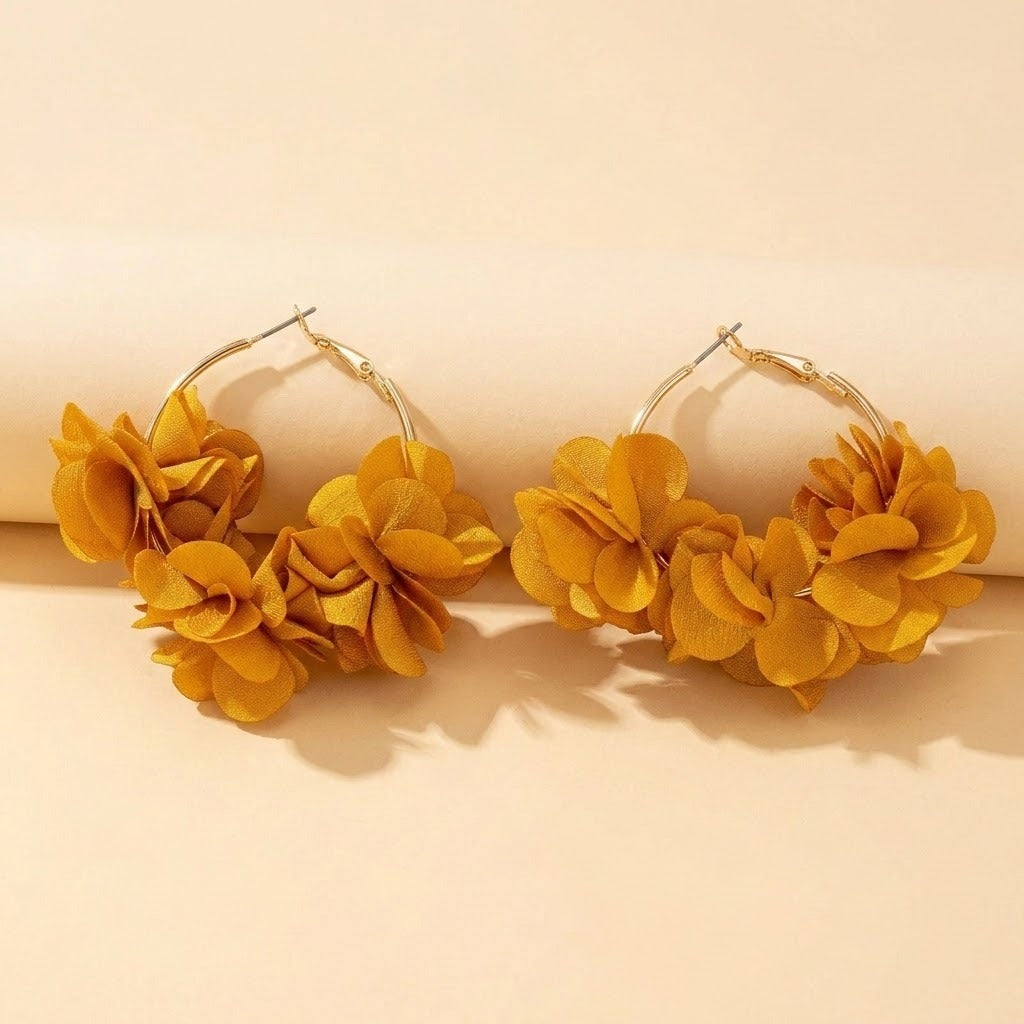 A pair of large gold-tone hoop earrings adorned with clusters of layered mustard yellow fabric flowers, displayed on a warm beige background.