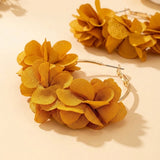 Gold-tone hoop earrings adorned with layered, flower-like fabric petals in a warm mustard yellow color, displayed on a light beige surface.