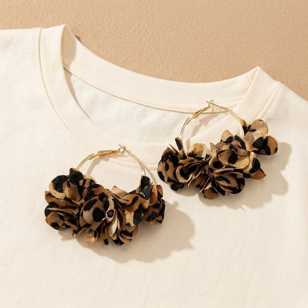 A pair of gold hoop earrings with layered leopard-print fabric flower clusters, displayed on a white t-shirt against a beige background.