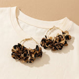 A pair of gold hoop earrings with layered leopard-print fabric flower clusters, displayed on a white t-shirt against a beige background.