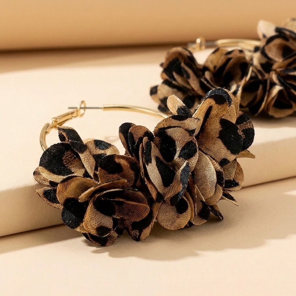 Tan and black leopard-print fabric flowers cover a gold-tone hoop earring on cream display blocks, with another earring behind.
