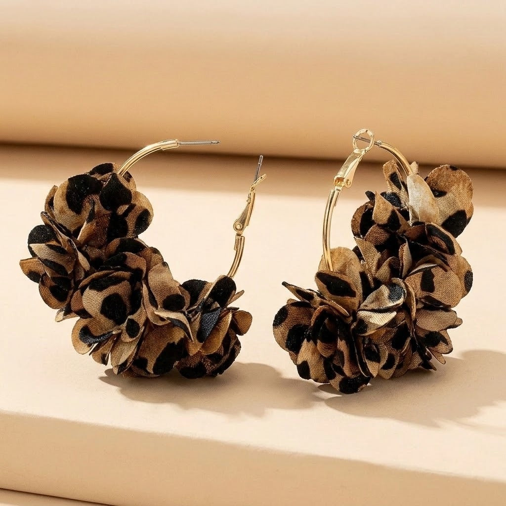 Two gold-tone hoop earrings with tan and black leopard-print fabric flowers stand upright on a cream platform, showing open clasps.