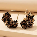 Two gold-tone hoop earrings with tan and black leopard-print fabric flowers stand upright on a cream platform, showing open clasps.