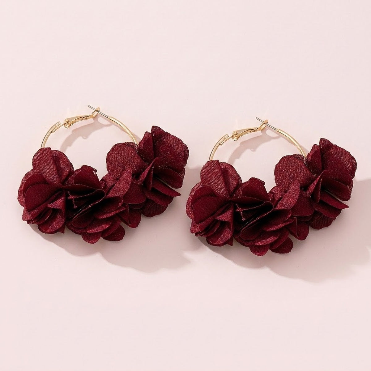 Two gold-tone hoop earrings with burgundy red fabric flower clusters lie side by side on a pale pink background.