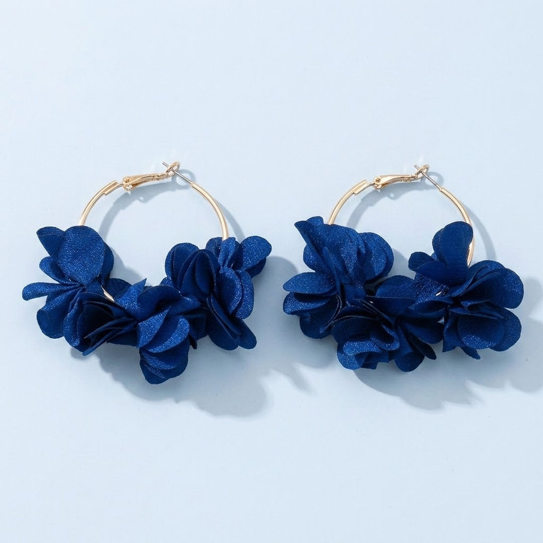 Two gold-tone hoop earrings decorated with deep blue fabric flowers lie on a pale blue background, casting soft shadows.