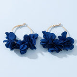 Two gold-tone hoop earrings decorated with deep blue fabric flowers lie on a pale blue background, casting soft shadows.