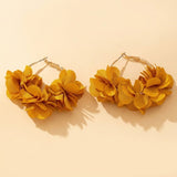 Two gold-tone hoop earrings with mustard yellow fabric flower clusters rest on a pale cream background, casting distinct shadows.
