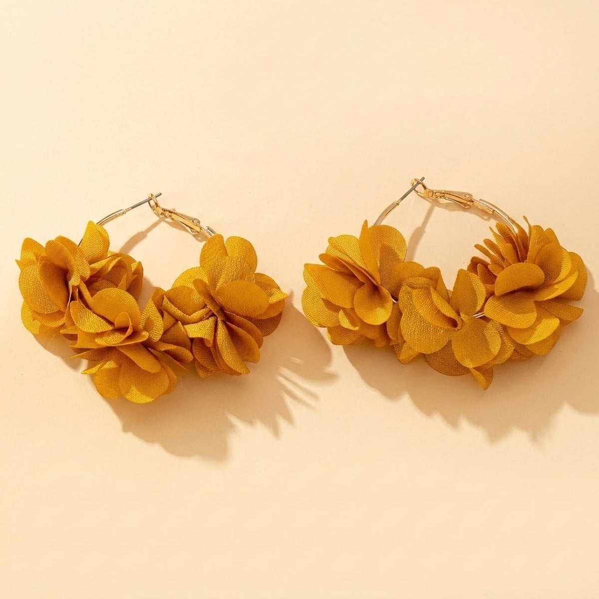 Two gold-tone hoop earrings with mustard yellow fabric flower clusters rest on a pale cream background, casting distinct shadows.