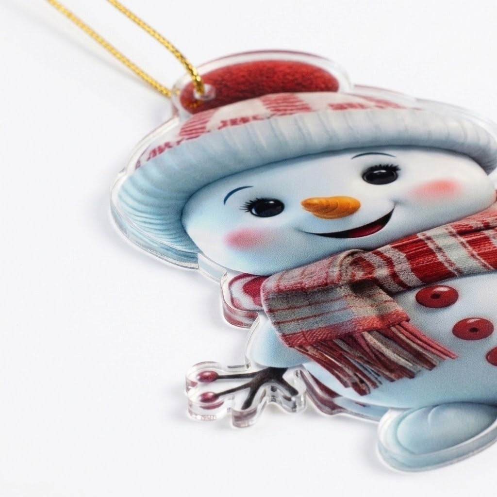 A flat acrylic snowman Christmas ornament with a clear edge, pale blue hat, and red plaid scarf rests against a white background.